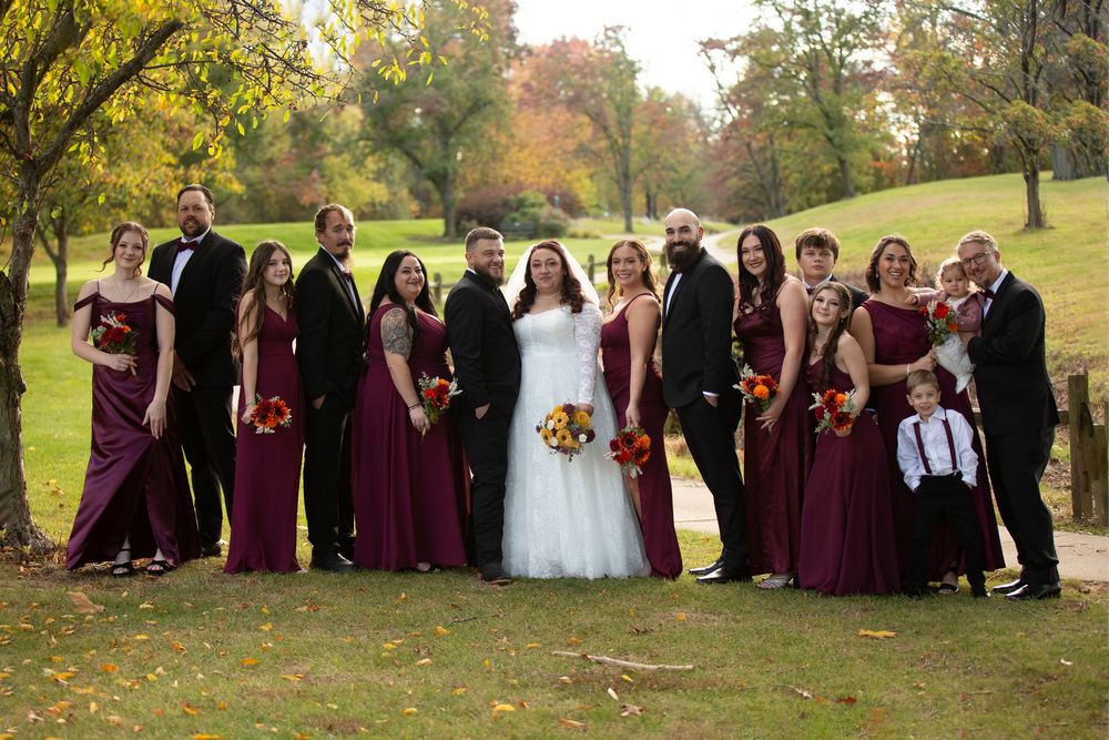 Wedding party in burgundy dresses and black suits posing outdoors on a lush green lawn with autumn foliage in the background.