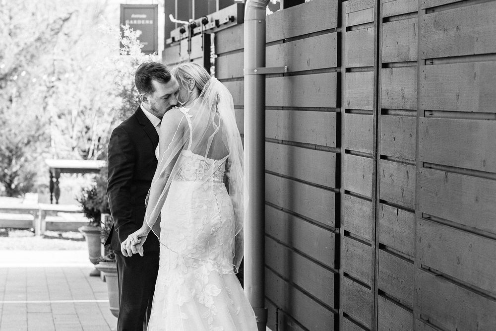 Black and white photo of bride and groom sharing a tender moment, leaning in close beside wooden lockers outdoors.