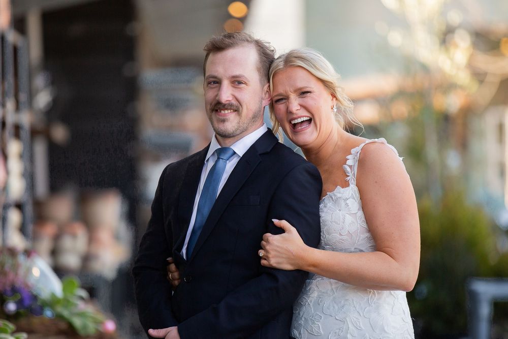 Happy bride and groom laughing together outdoors on their wedding day, bride in lace gown, groom in navy suit with blue tie.