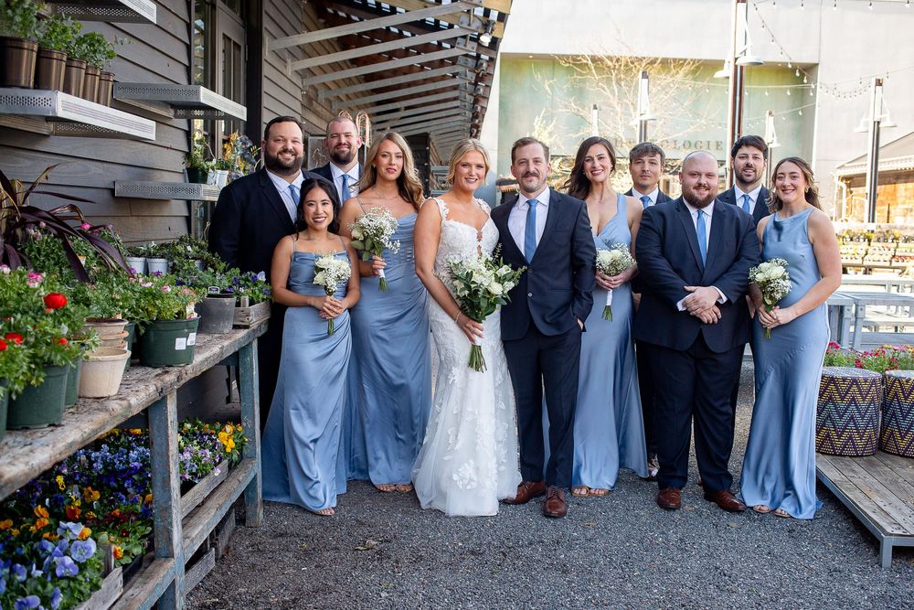Wedding party in blue gowns and suits posing among potted flowers in a rustic greenhouse venue.