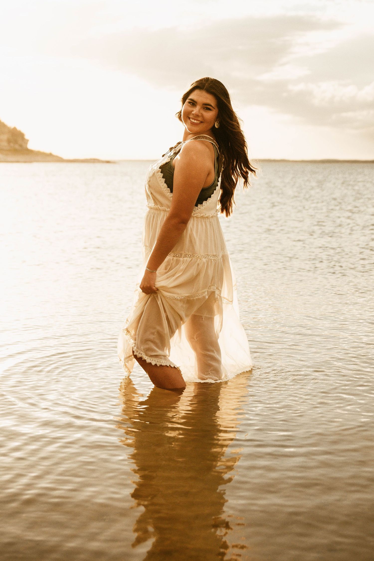 A person in a white dress wades through calm ocean waters at sunset creating a dreamy, ethereal scene.