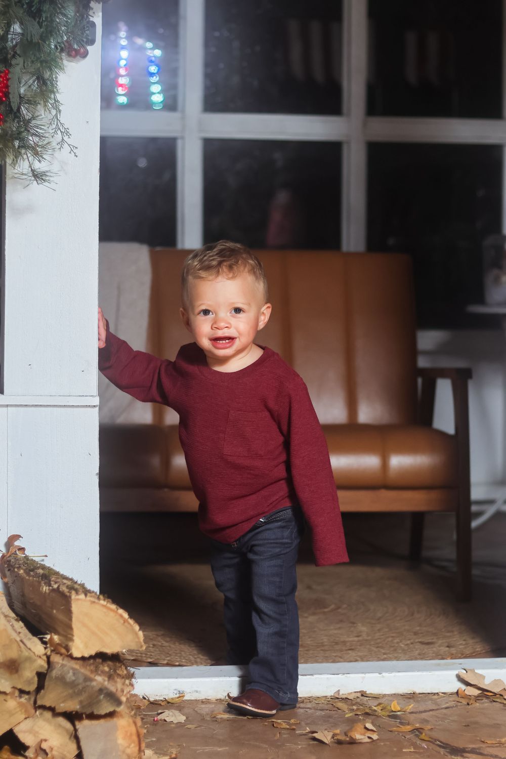 A small child in a burgundy sweater and jeans explores a wooden porch area with fallen autumn leaves.