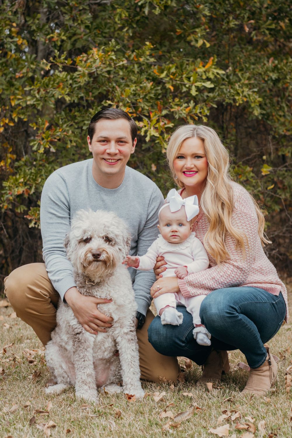 A family sits together outdoors in autumn with their fluffy white dog and baby on fallen leaves.