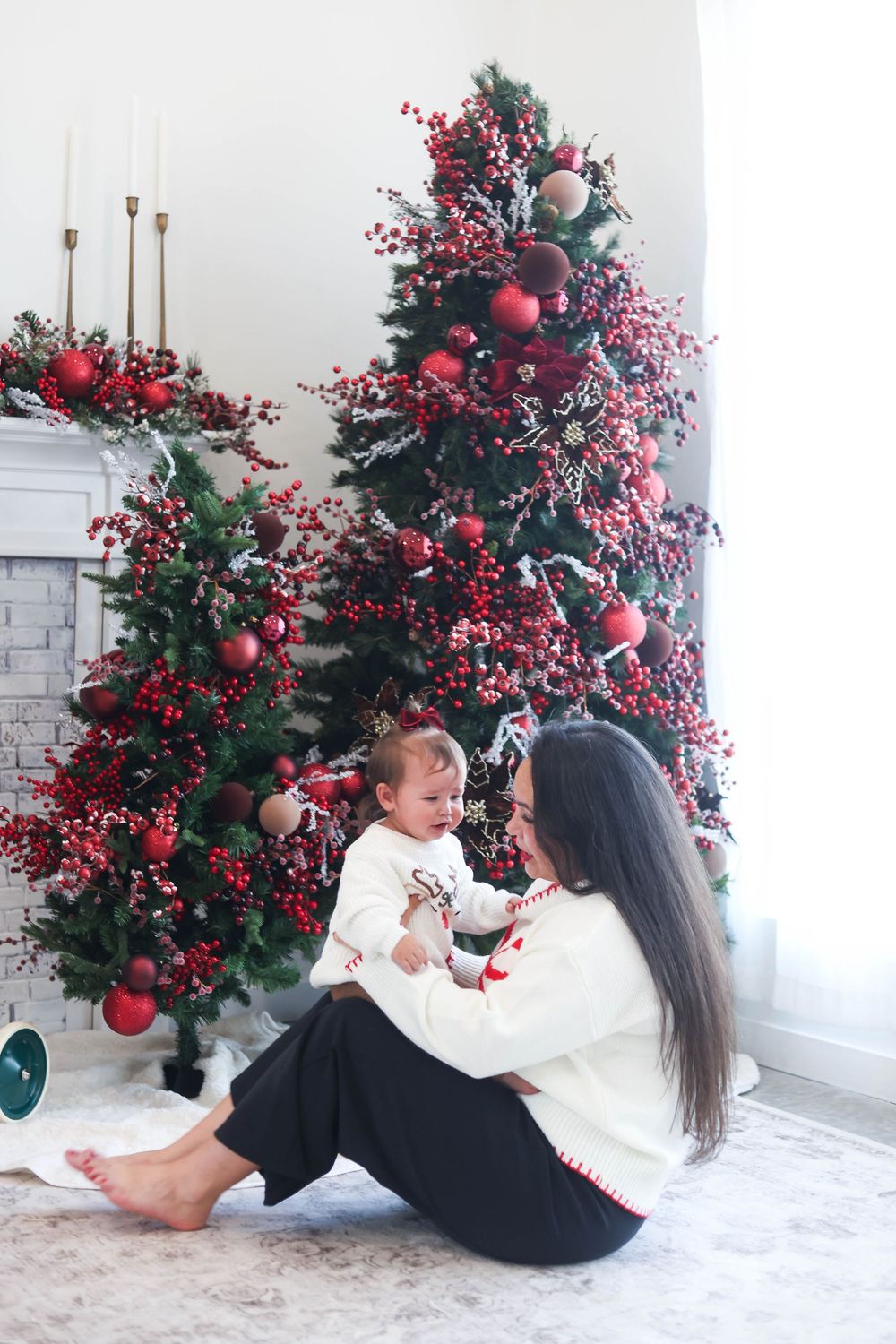 Mother and child in matching white sweaters share playful moments by a red and green decorated Christmas tree.