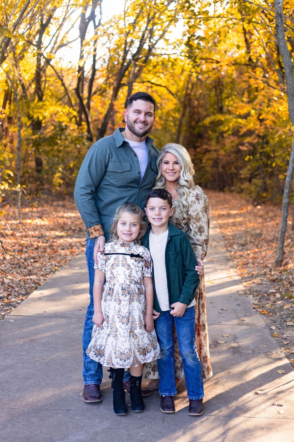 Family poses together on a sunlit autumn path surrounded by golden yellow trees and fallen leaves.