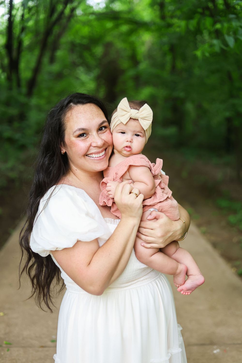 A woman in a white dress holds and plays with a baby outdoors in a wooded setting.