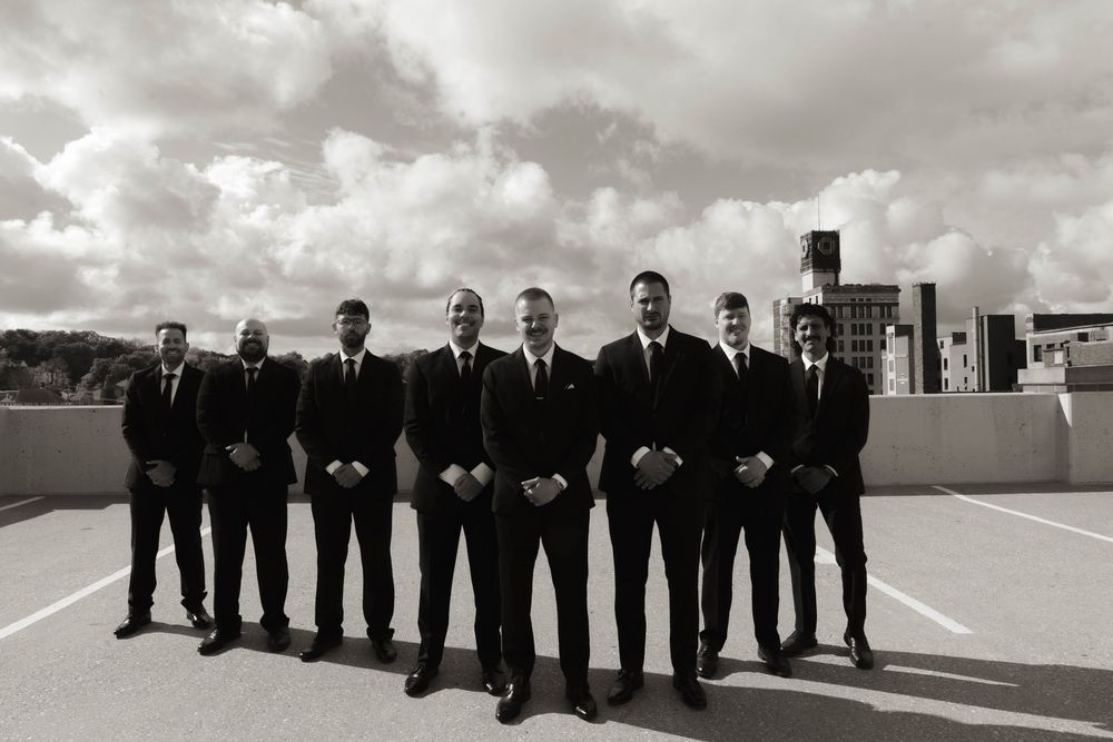 Groomsmen in black suits stand in a line on a rooftop with dramatic cloudy skies.