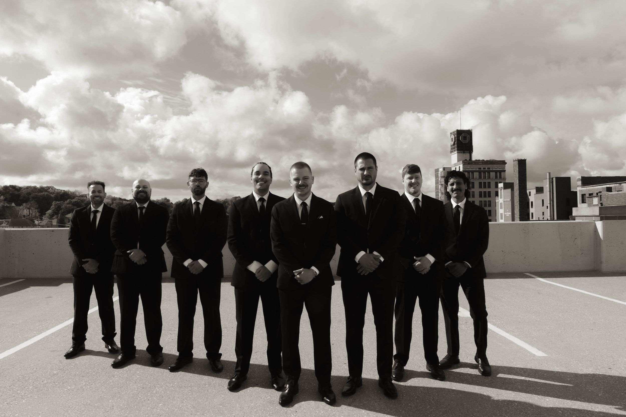 Groomsmen in black suits stand in a line on a rooftop with dramatic cloudy skies.