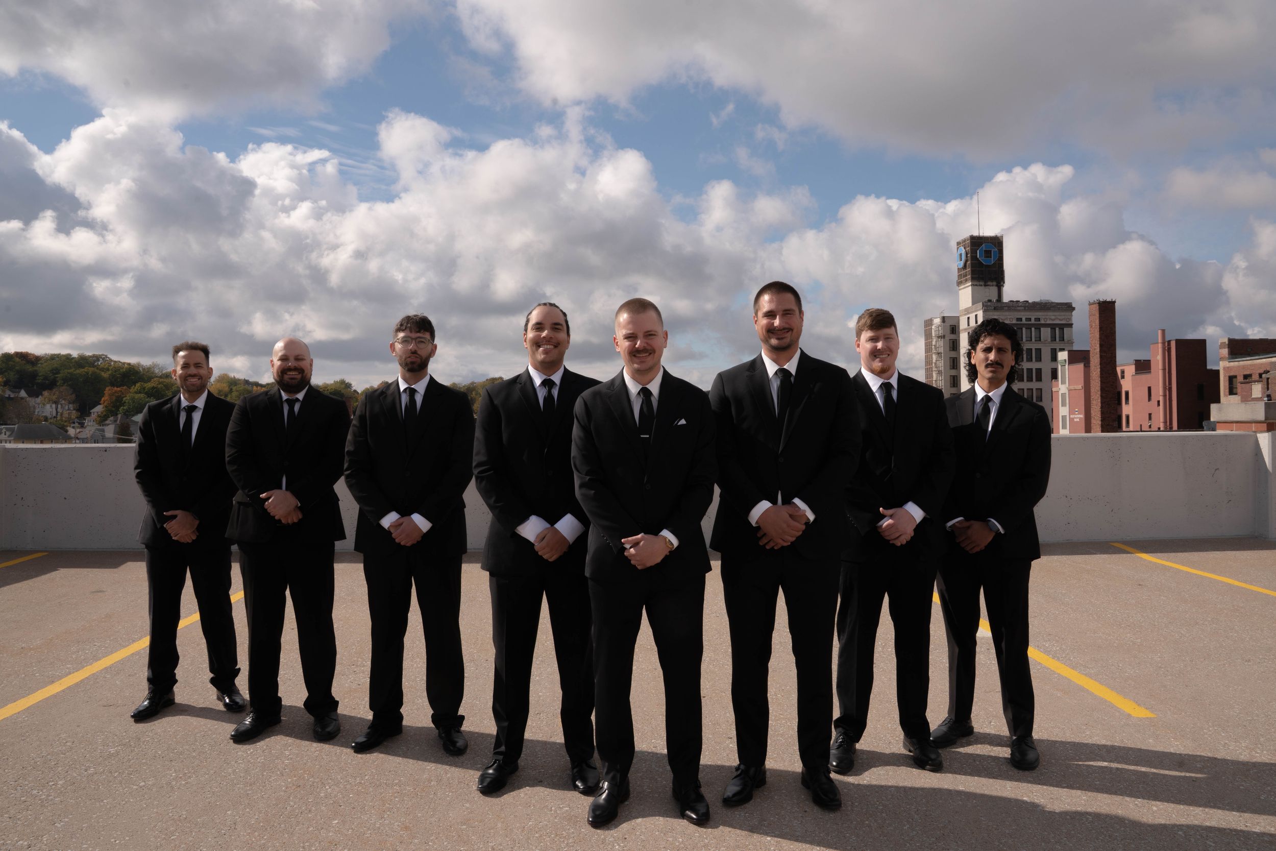Wedding party in matching black suits poses together on a rooftop with city buildings in background.