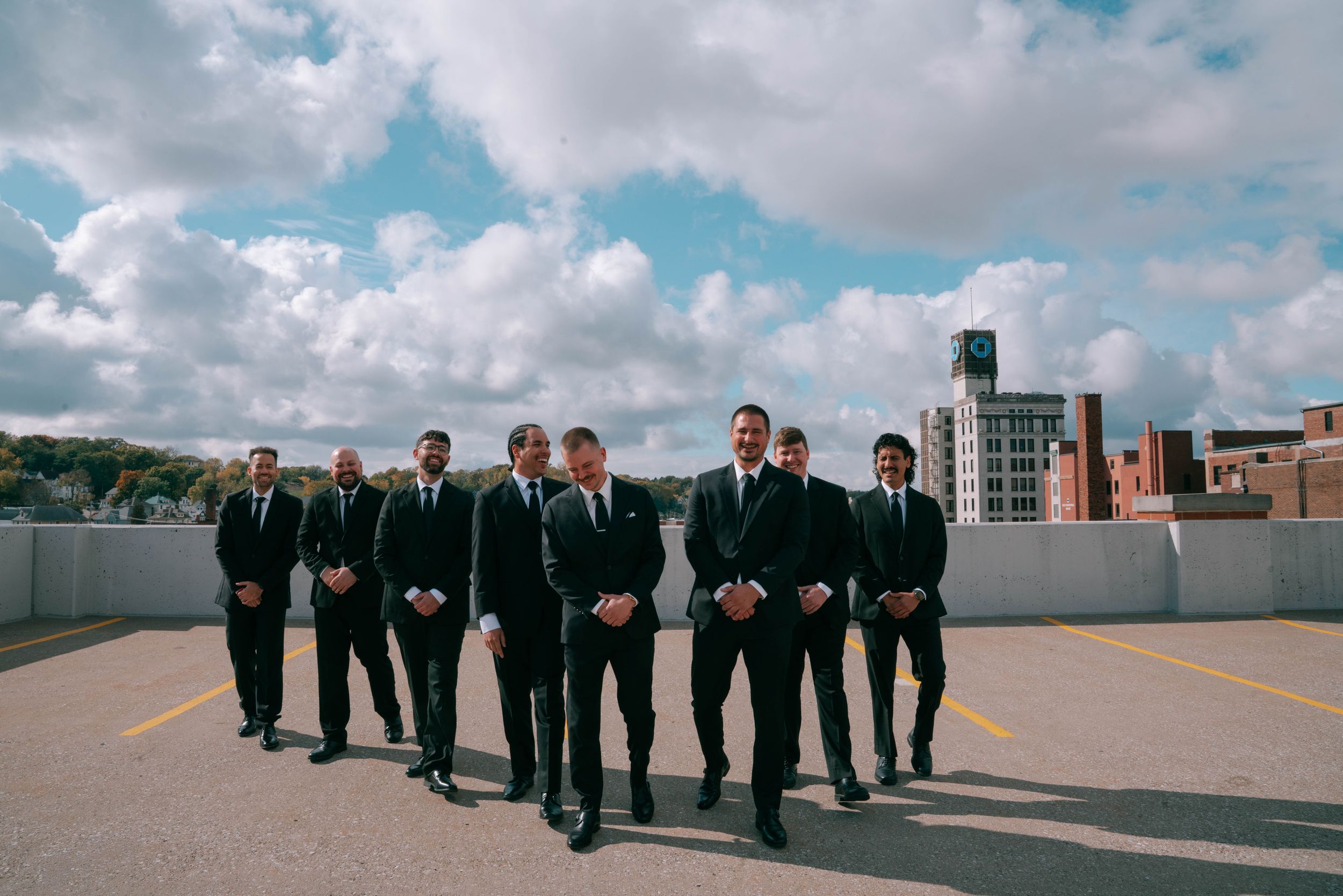 Group of groomsmen walking in formation wearing black suits on an urban rooftop.