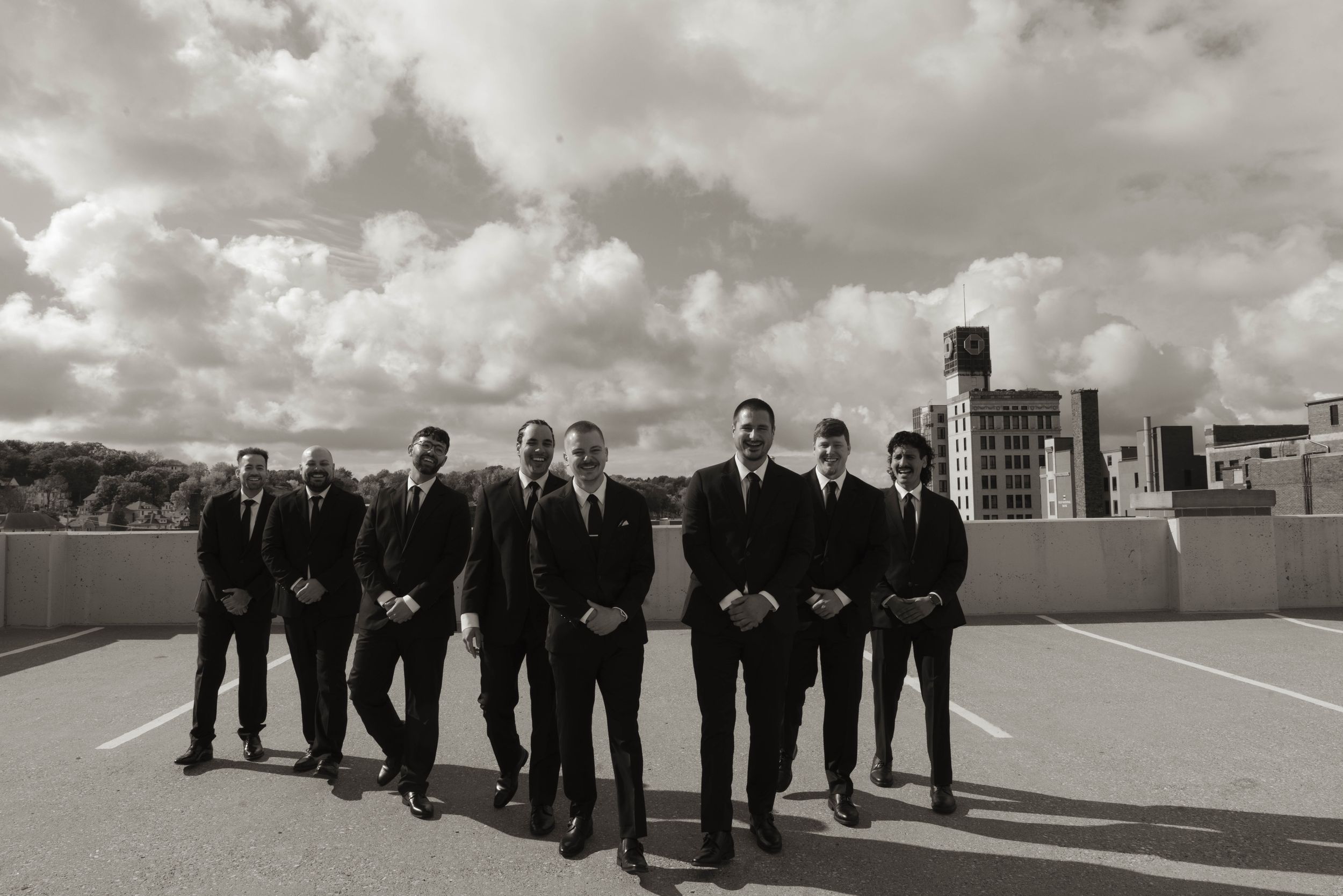 Wedding party in formal attire stands lined up against dramatic sky on building rooftop.