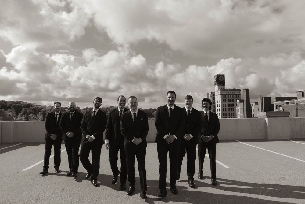 Wedding party in formal attire stands lined up against dramatic sky on building rooftop.