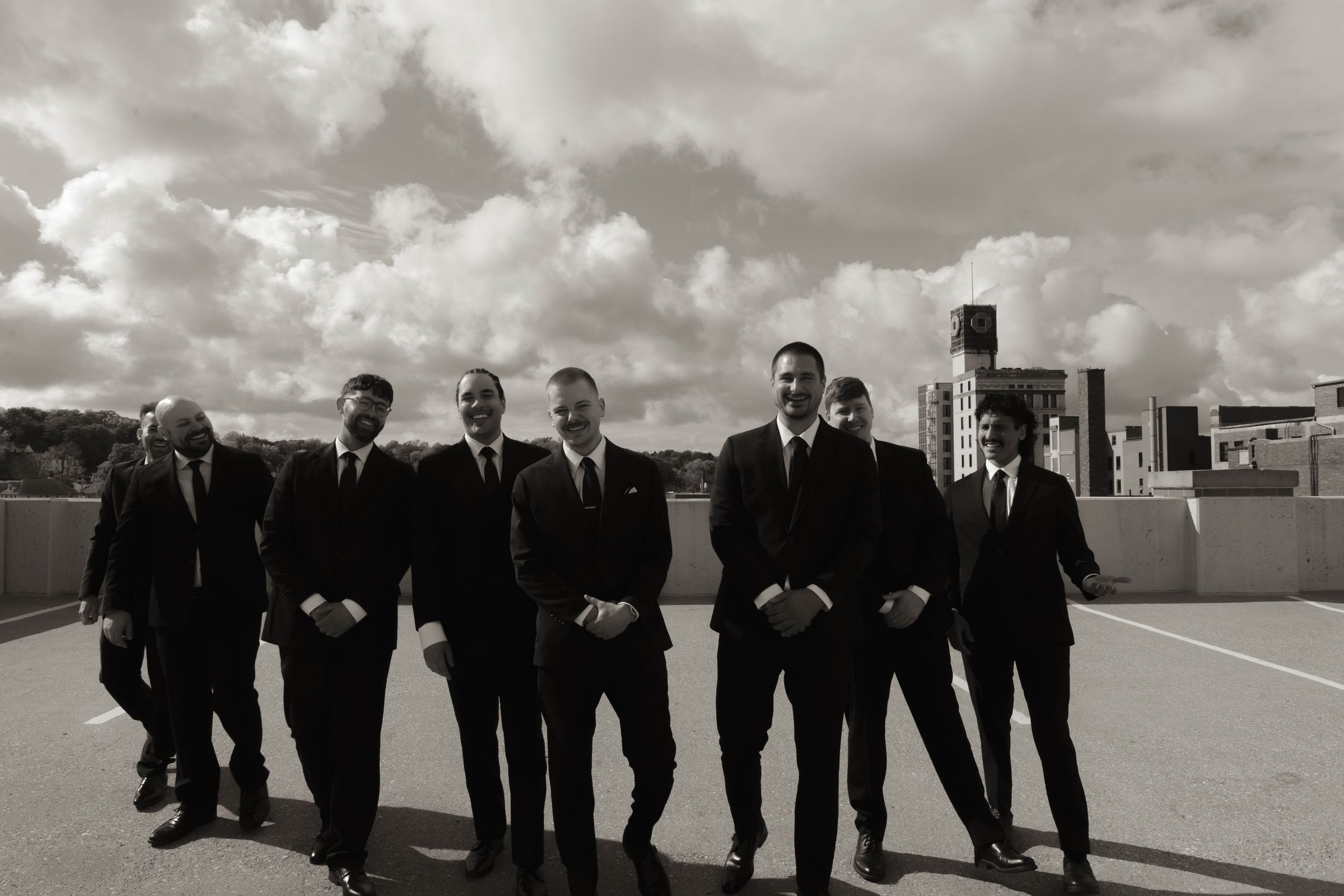 Groomsmen in dark suits pose casually on a rooftop with urban architecture in the background.