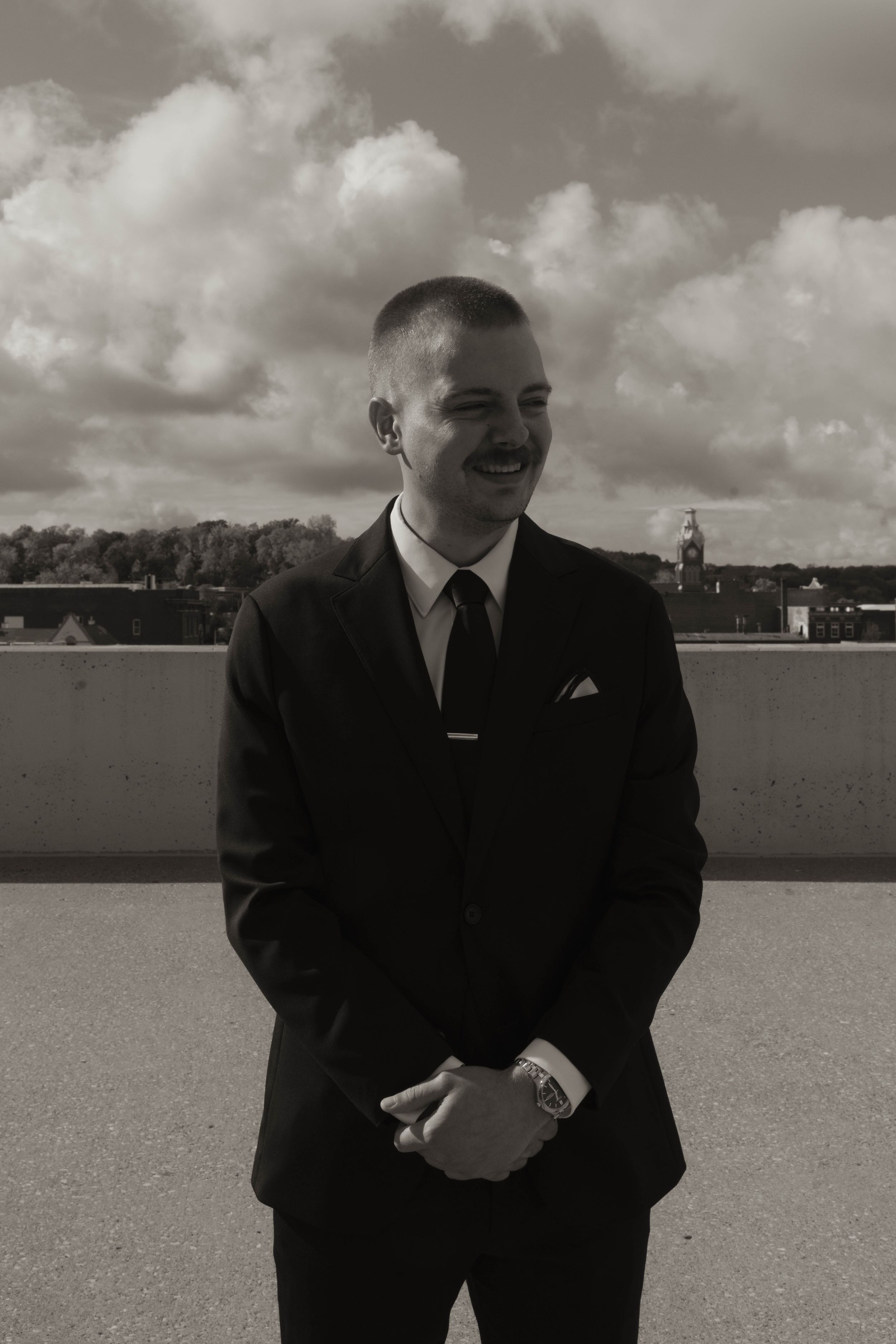 A person in a classic black suit smiles while standing on a rooftop under cloudy skies.