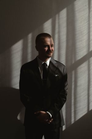 A person in a dark suit stands in dramatic window light casting shadows on the wall.
