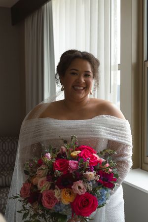 Person in white wedding dress holding vibrant pink and red floral bouquet by window.