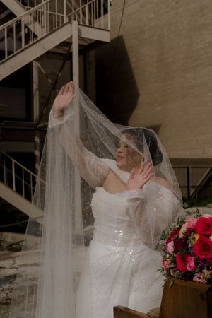 A bride in a sparkling dress plays with her veil near a metal staircase.