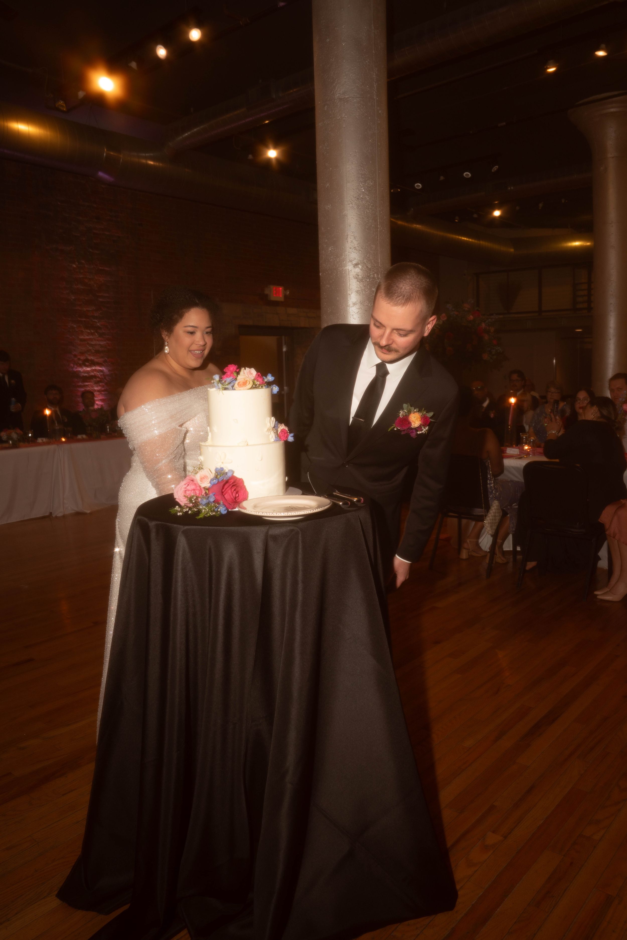 A couple cuts their white wedding cake together at an elegant reception with black tablecloths and dim lighting.