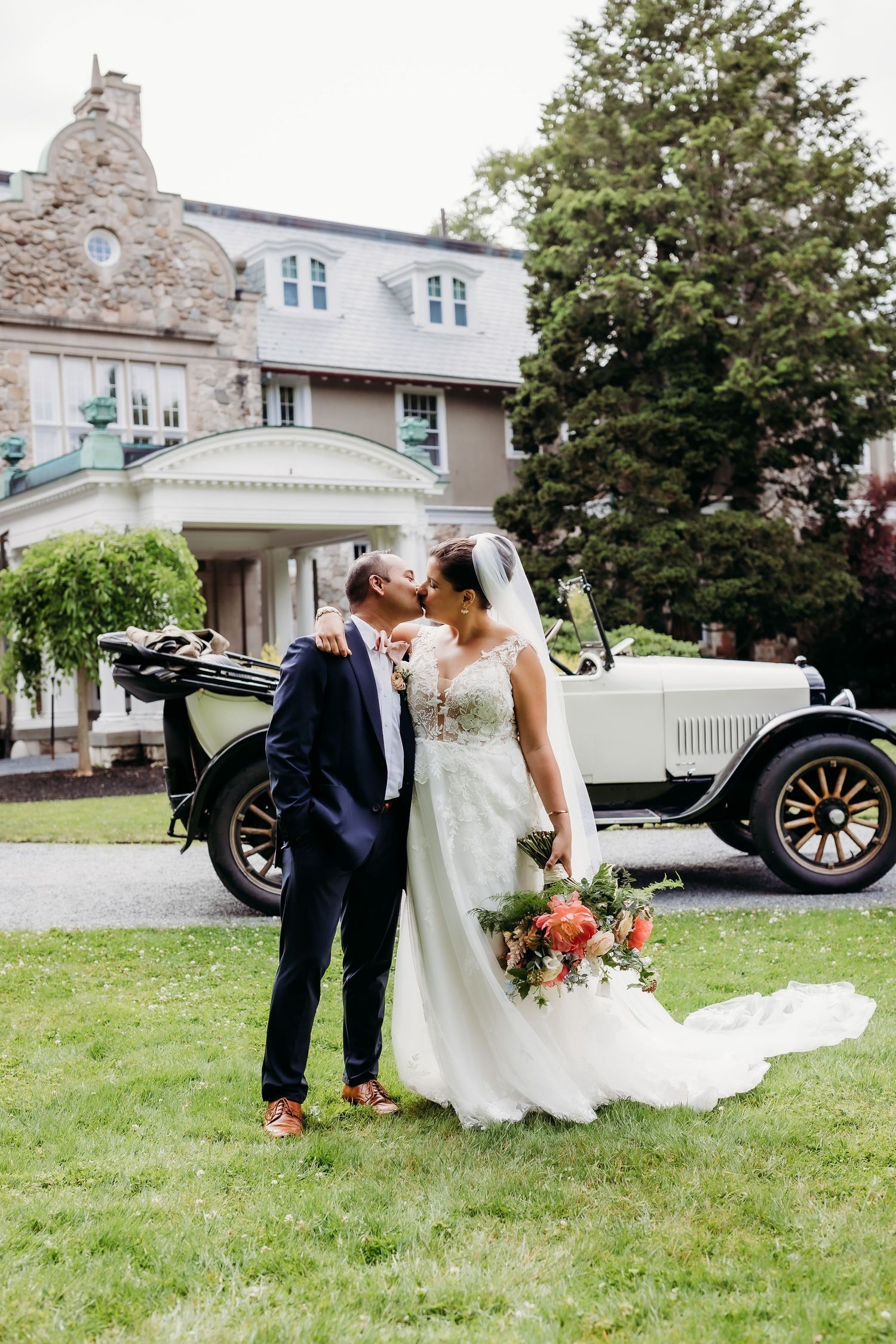 Wedding couple poses near vintage car outside historic stone building.