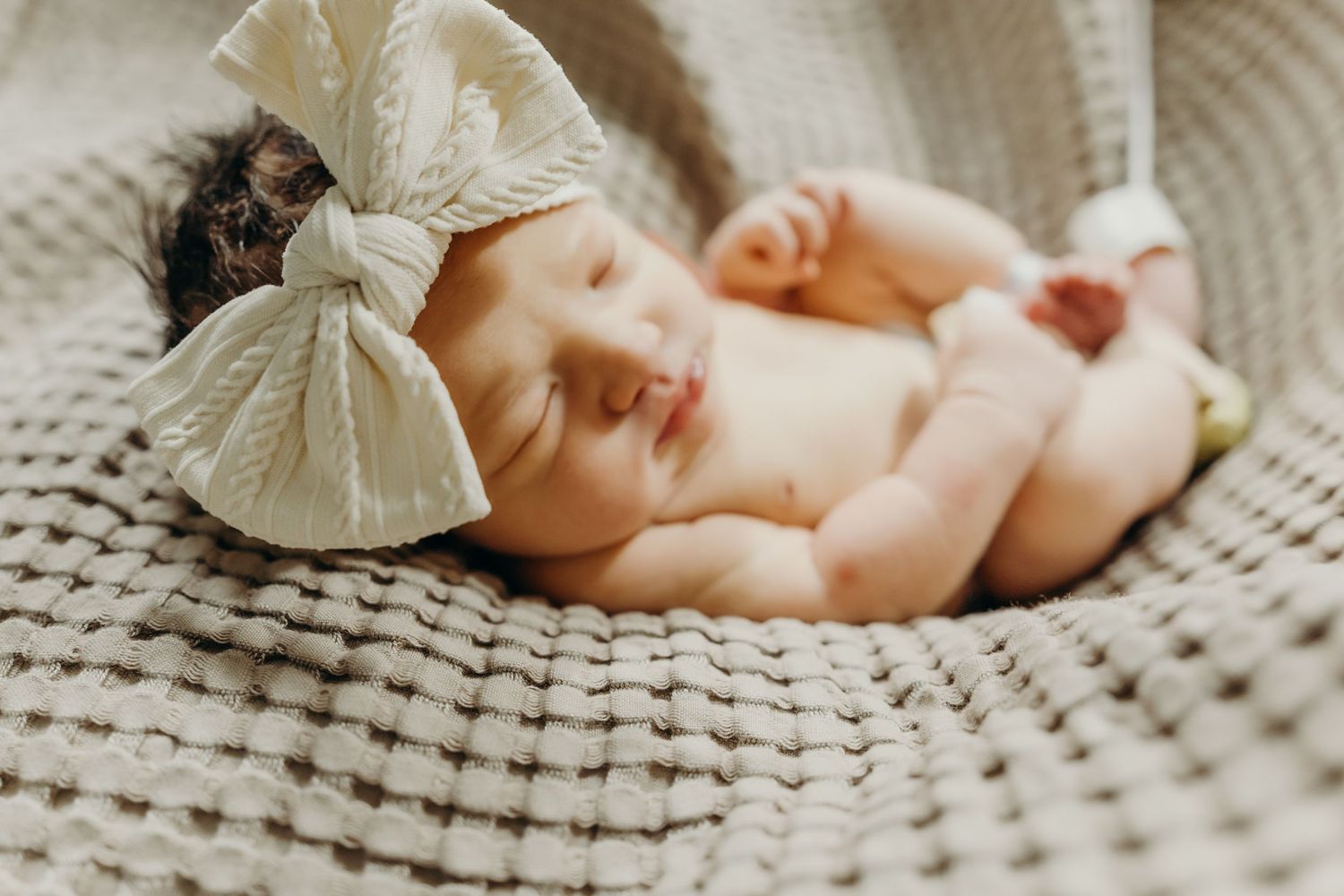 Sleeping newborn wearing large cream bow on textured knit blanket.