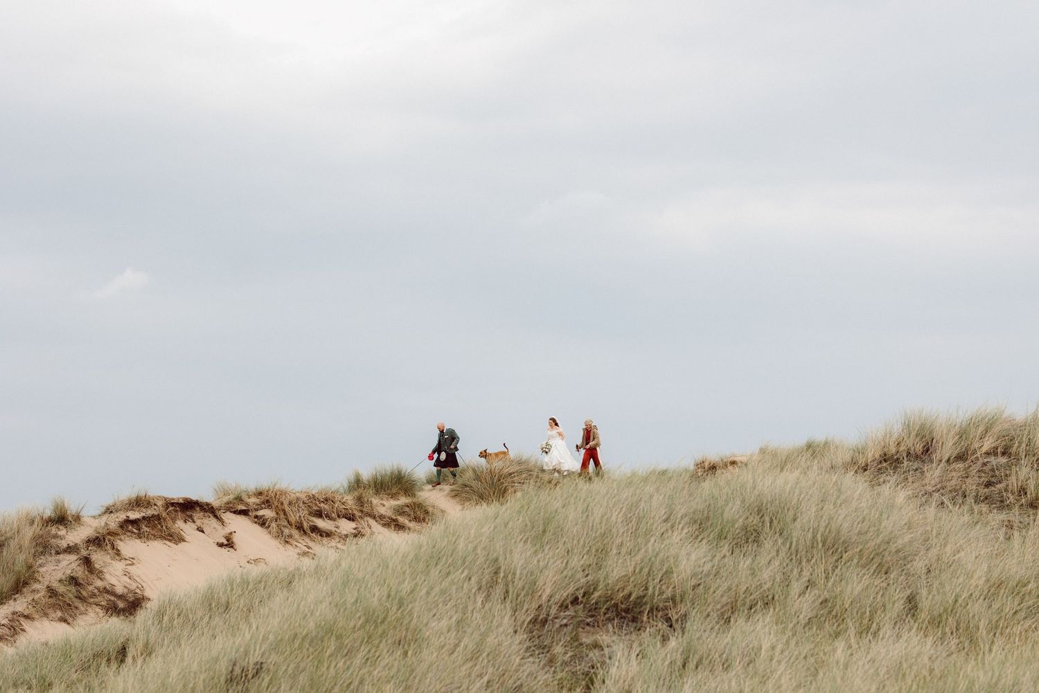 People walking along the crest of a sandy beach dune covered in tall grass against a cloudy sky.