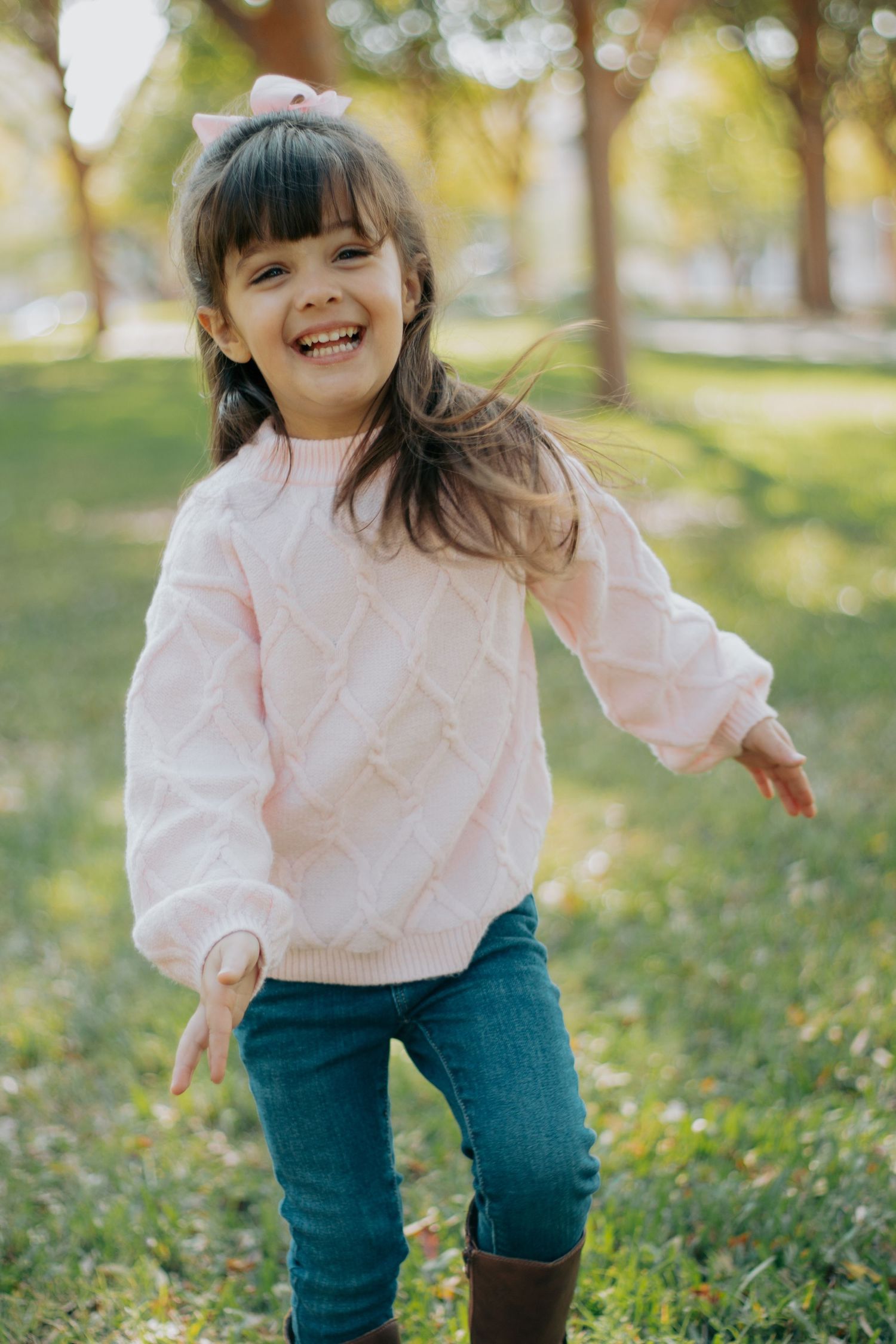A child in a pink sweater and jeans plays outdoors on a sunny autumn day in a park setting.