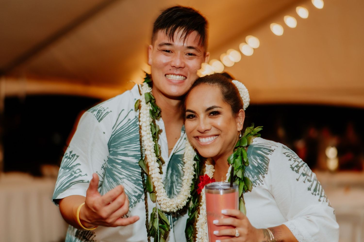 Reception celebration with couple wearing traditional Hawaiian leis under tent lighting.
