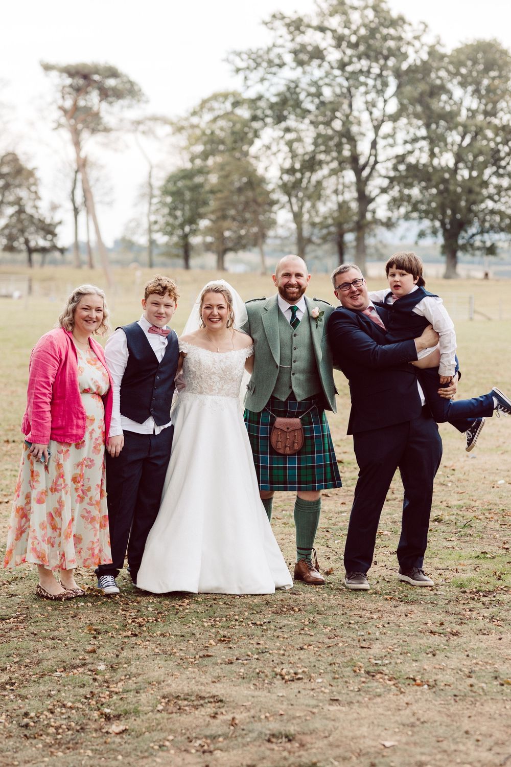 A wedding party poses playfully in a line on a grassy field during an autumn outdoor celebration.