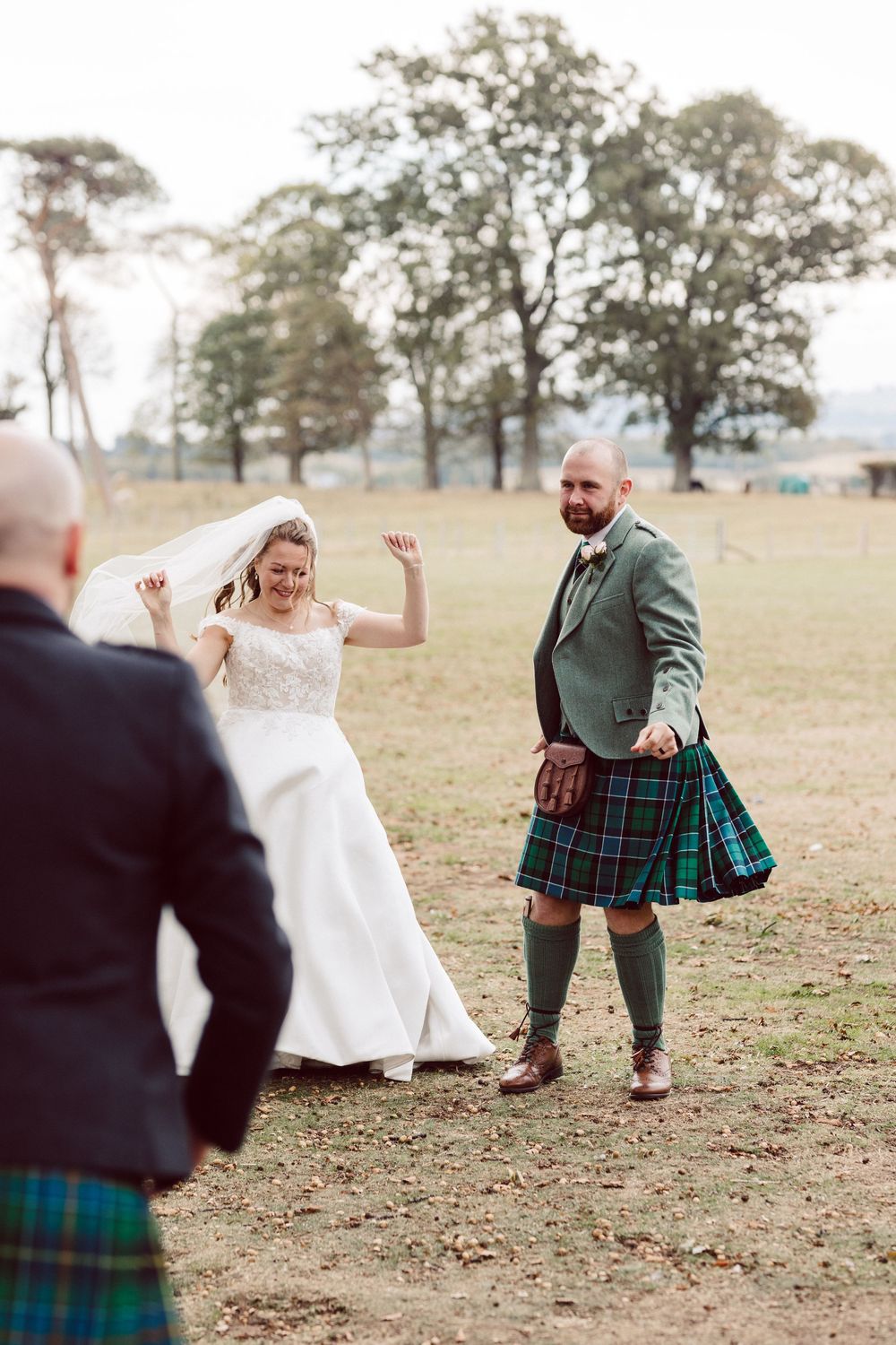 Playful couple in wedding attire and traditional Scottish kilt dancing together in an open field at sunset.