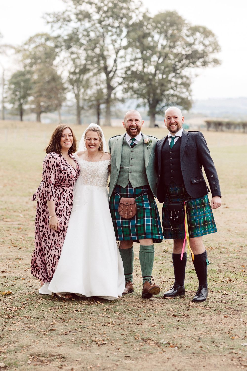 Wedding party wearing traditional Scottish kilts and formal attire poses together in an outdoor field setting.