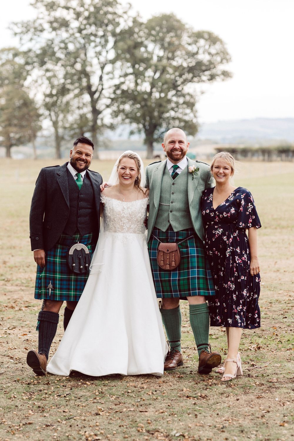 Wedding party wearing traditional Scottish kilts and formal attire pose together on autumn grounds.