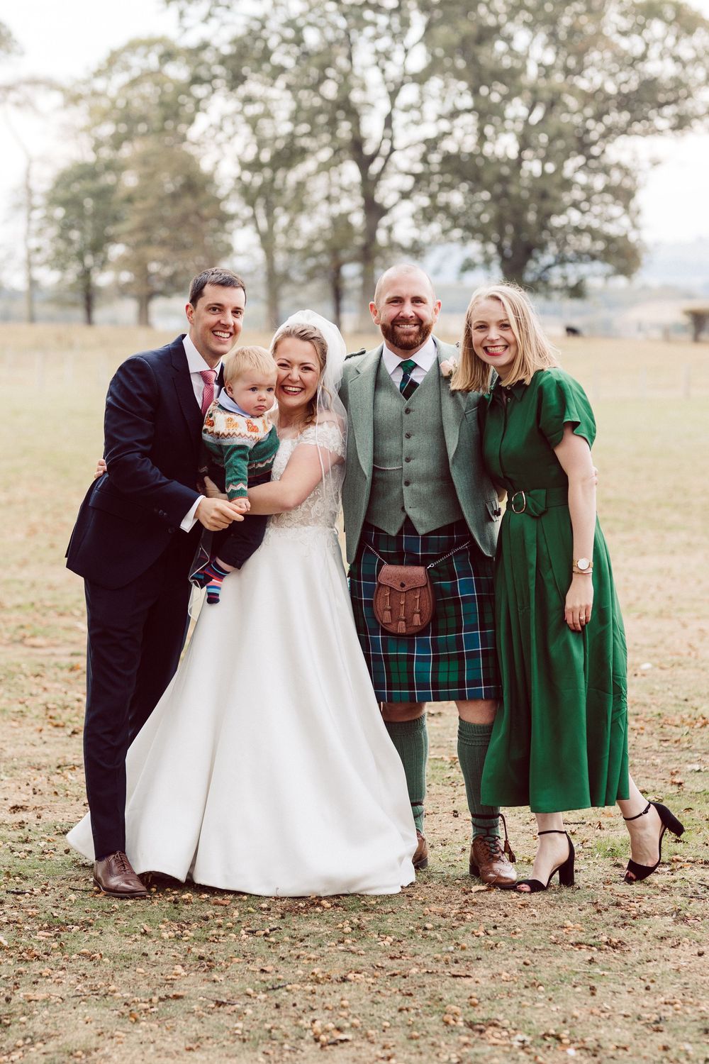 Wedding party in formal attire poses together outdoors on autumn day with fallen leaves on the ground.