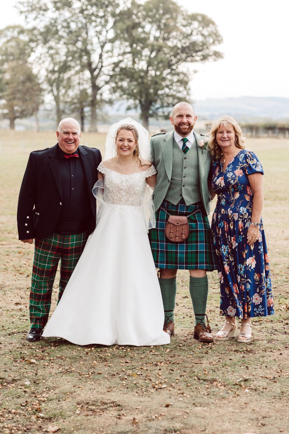 A wedding party poses outdoors wearing traditional Scottish kilts and formal attire in a natural setting.