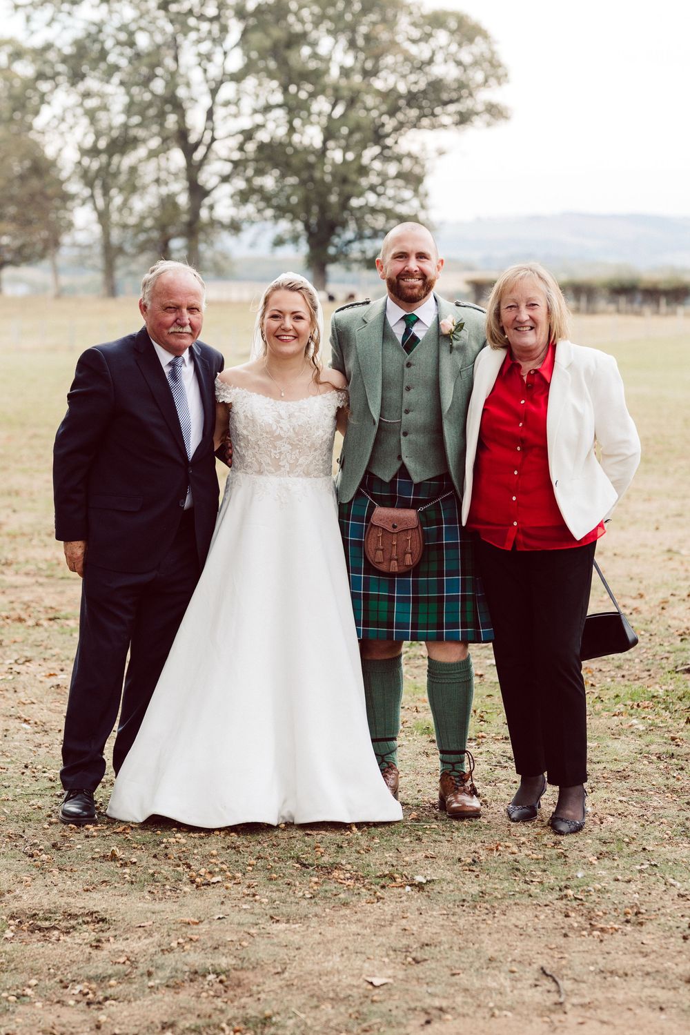 Wedding party stands together outdoors on autumn day wearing formal attire including kilts and traditional Scottish dress.