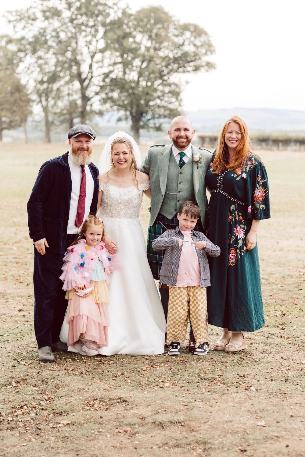 Wedding party poses together outdoors in formal attire on a fall day with dried grass and trees in background.