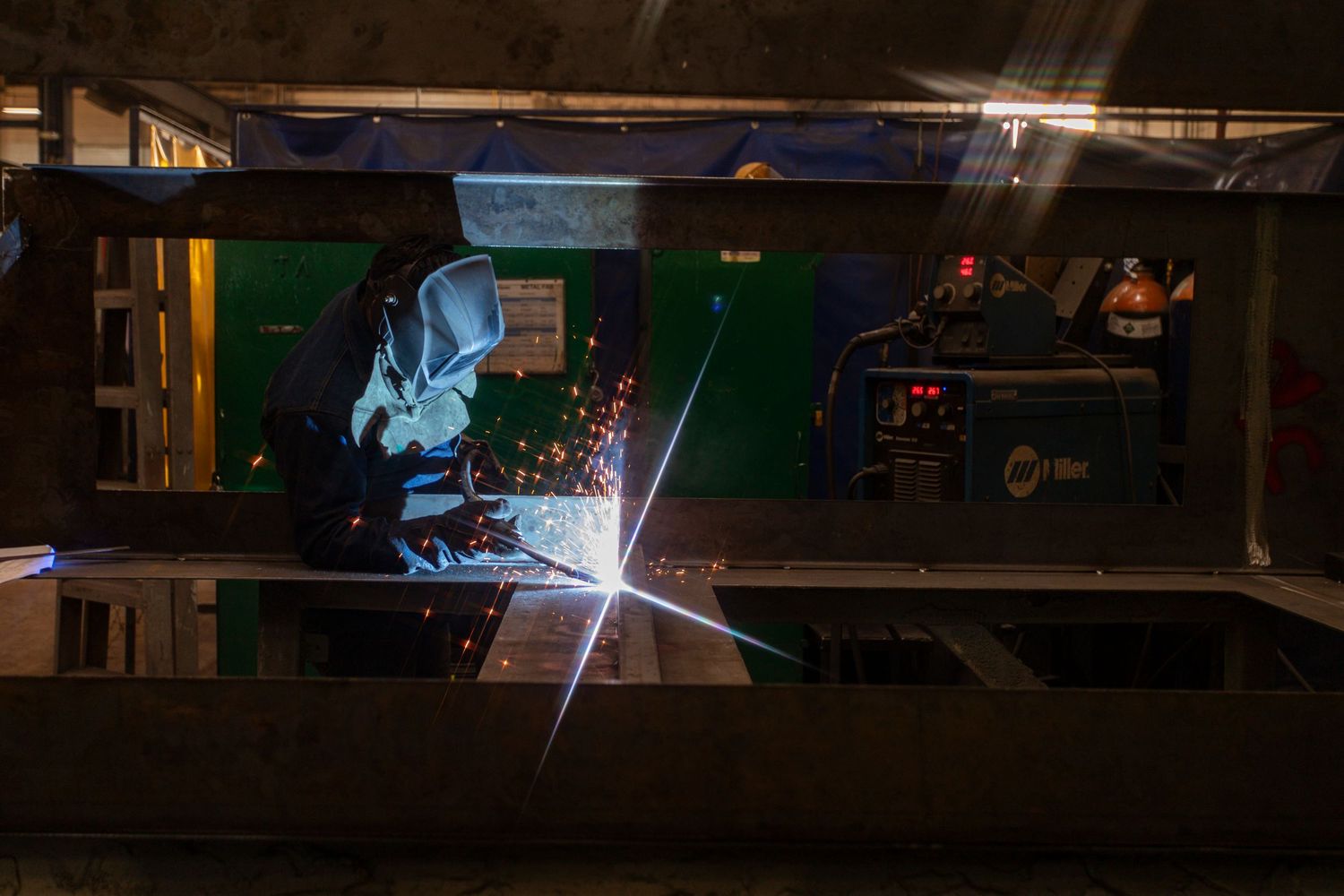 Sparks fly as a welder works in a dimly lit industrial workshop, creating a bright blue glow during metalwork.