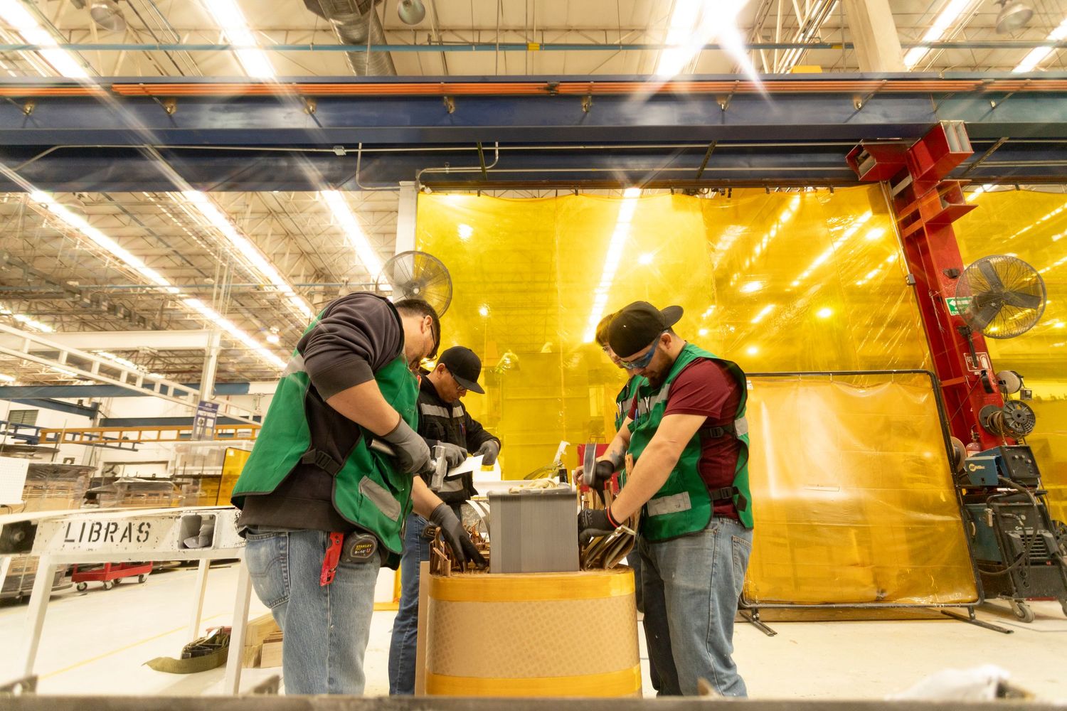 Two workers in green vests collaborate on a project in a brightly lit warehouse with yellow walls.