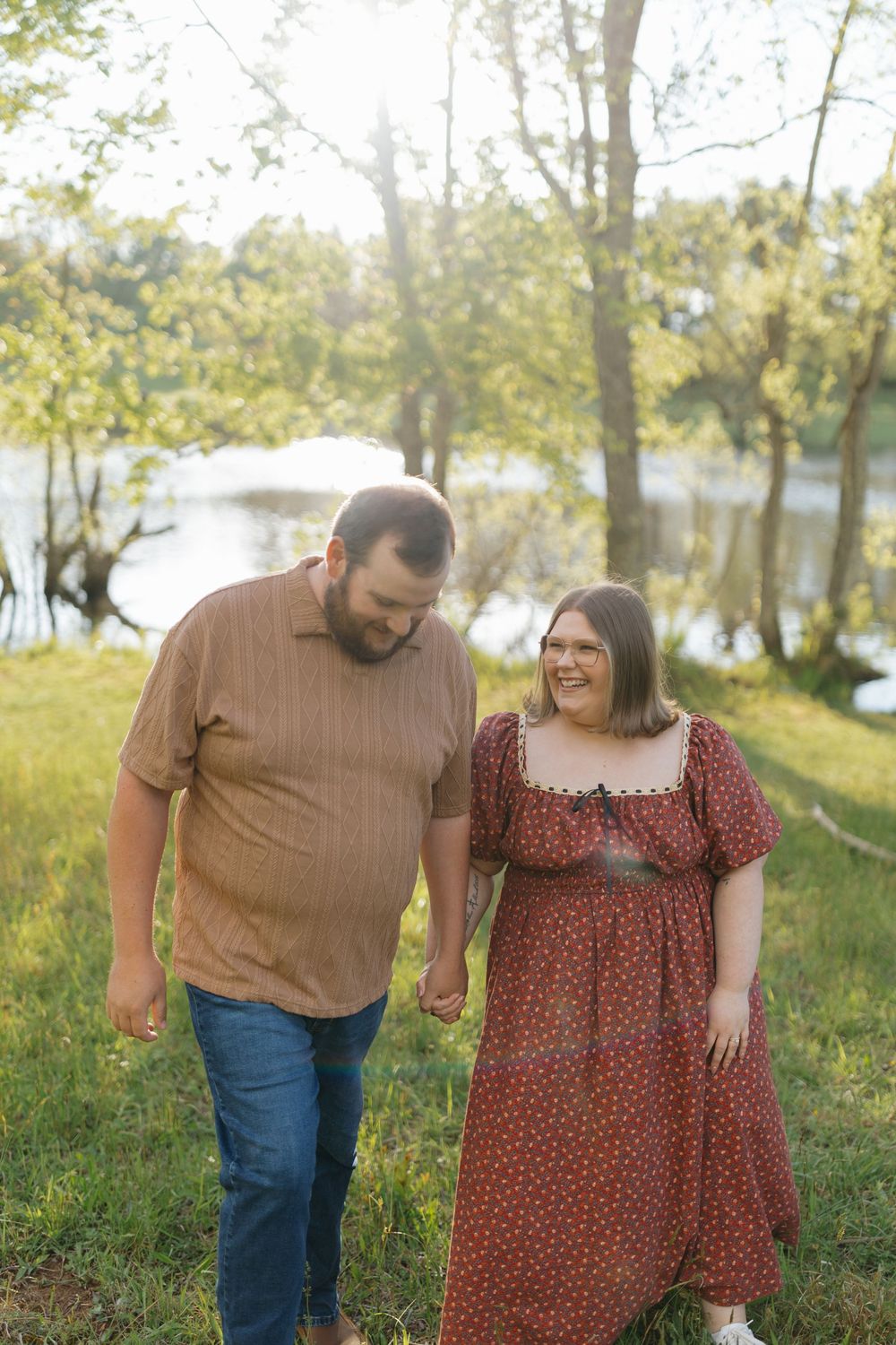 A couple shares a tender moment outdoors by a lake on a sunny day, with trees and grass in the background.