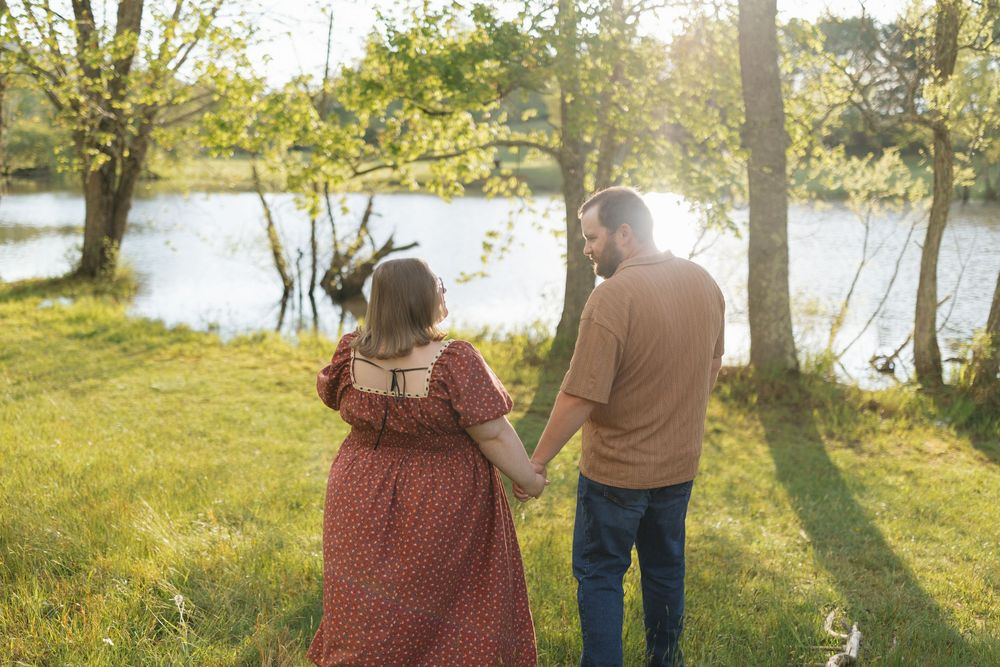 A couple walks hand in hand through a sunlit park with trees and a lake in the background during golden hour.