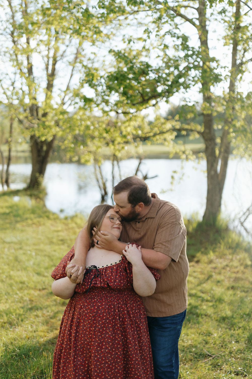 A couple laughs together during a romantic photoshoot in a scenic park setting with water and trees visible.