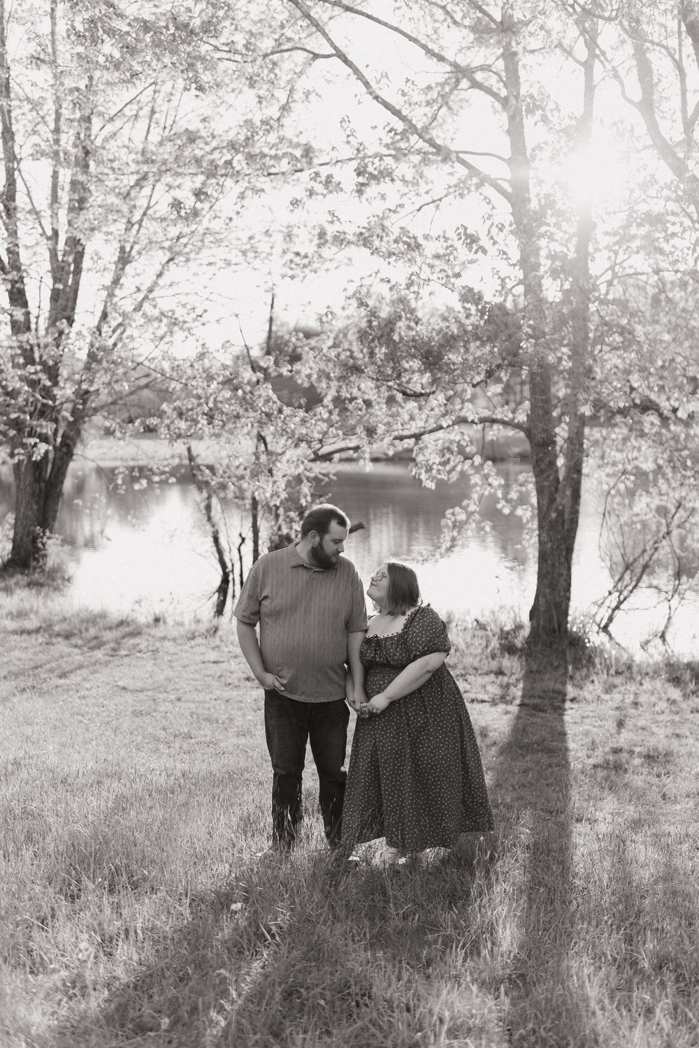 A romantic black and white photograph of a couple standing by a tranquil lake surrounded by trees.