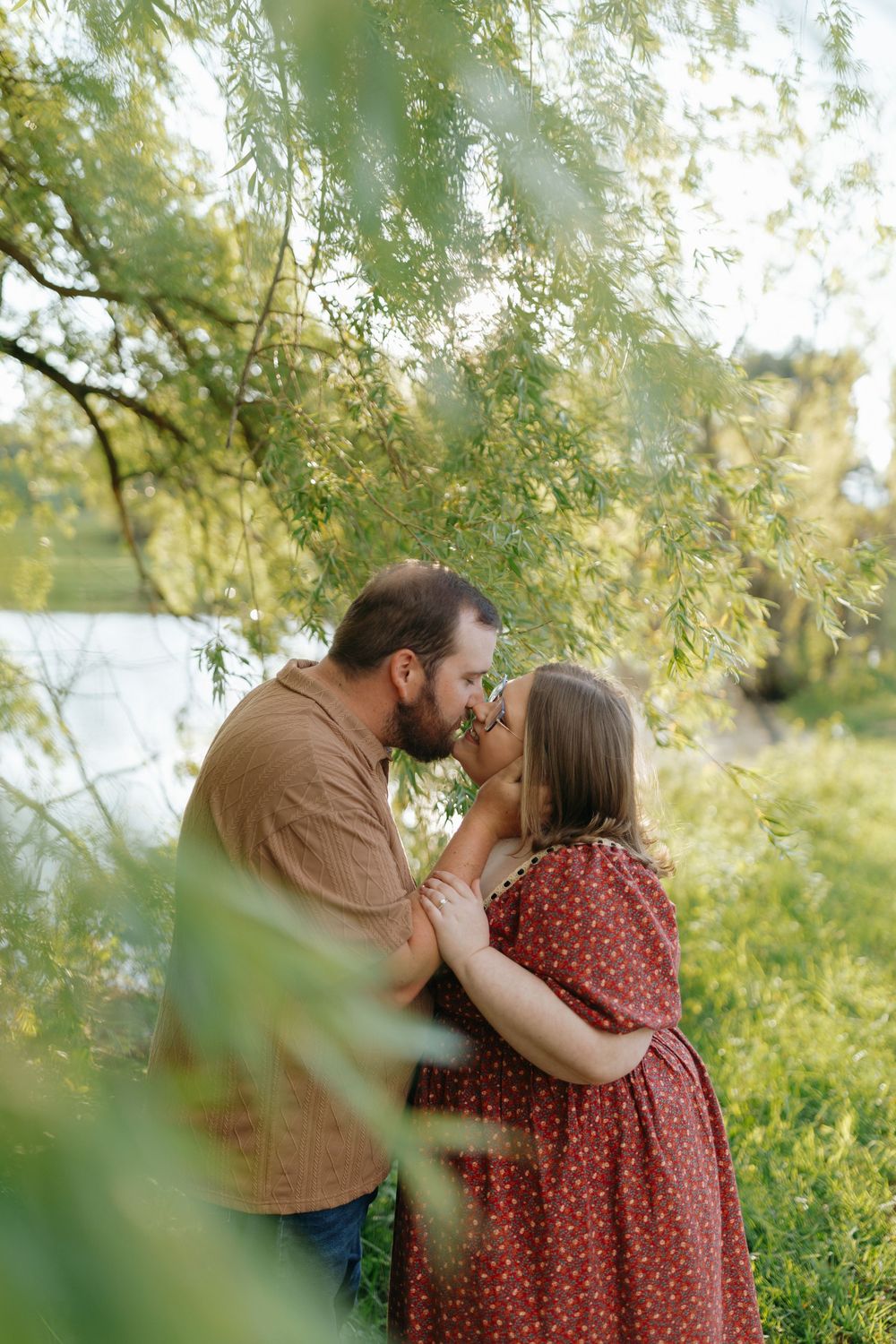 A couple shares an intimate moment under willow branches by a serene lake.