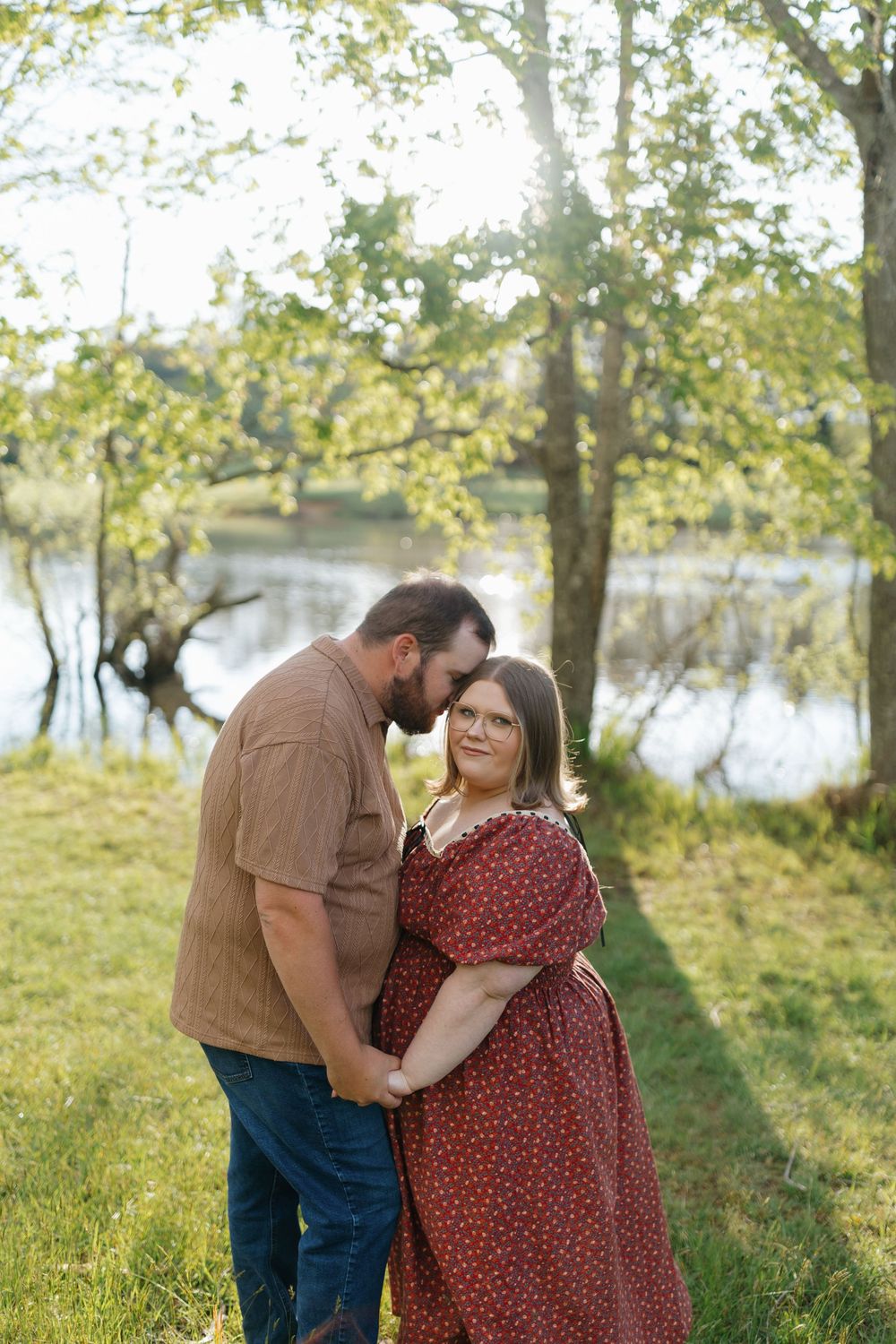 A couple shares tender moments in a sunlit meadow by a lake while wearing casual autumn attire.