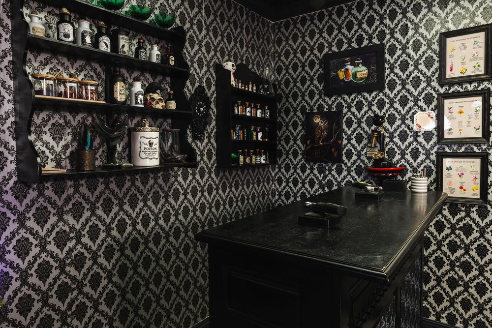 Dark bar interior with black and white geometric wallpaper, framed artwork, and glass bottle display shelving.