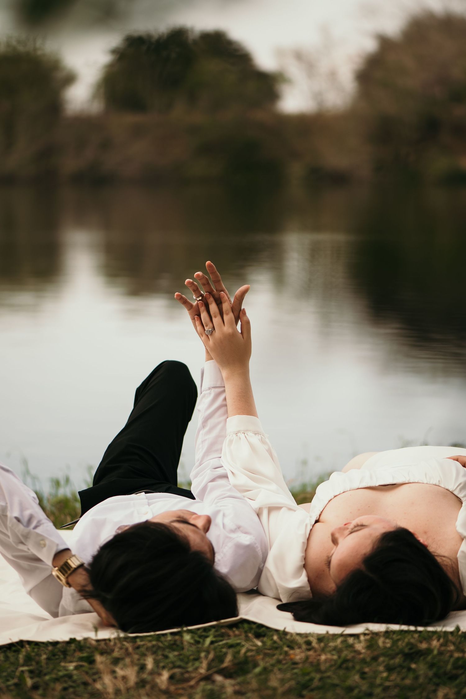 Two people in white clothing lie peacefully by a tranquil lake with their hands raised in a dreamy pose.
