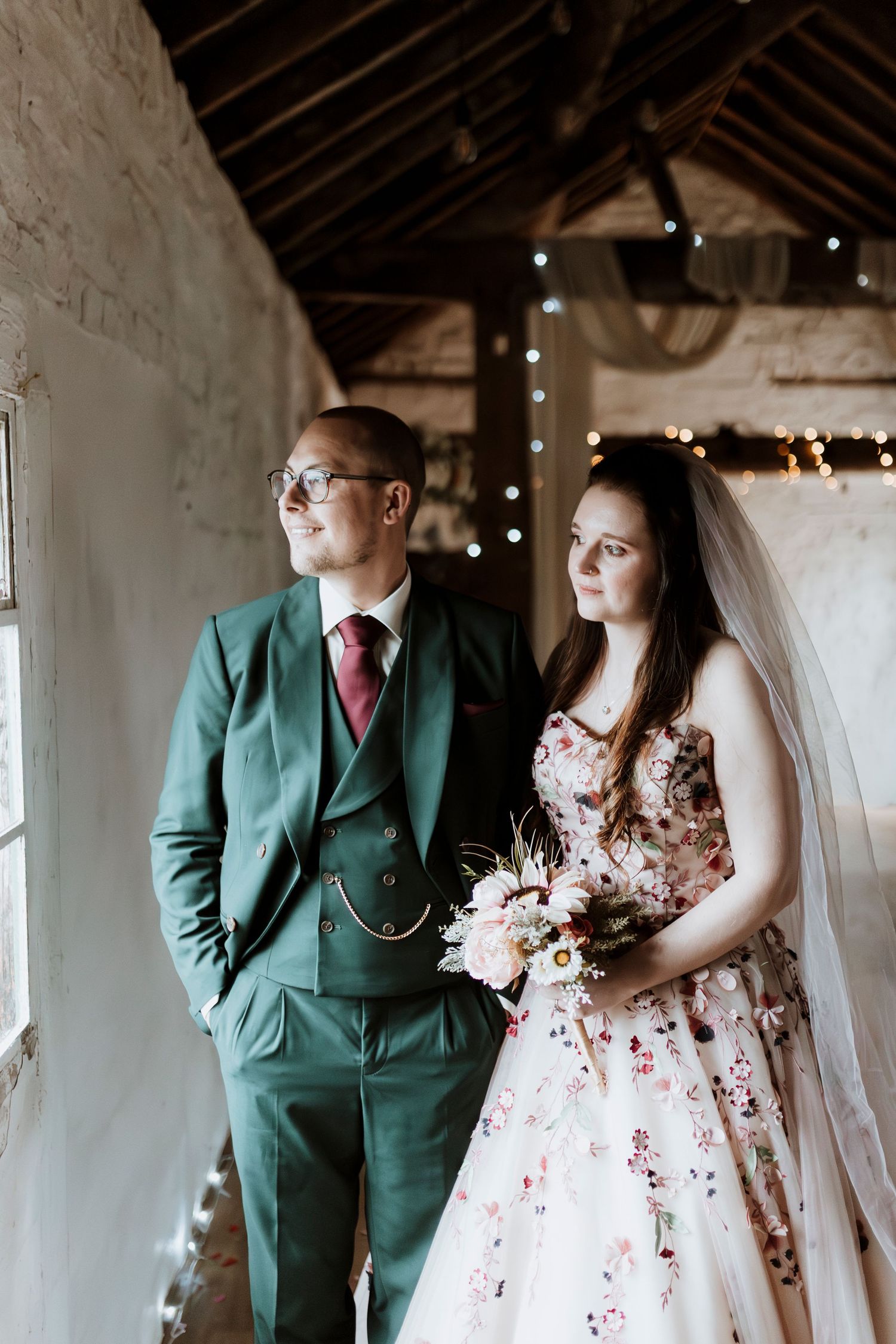 A couple in a rustic barn wedding setting with the groom wearing a green suit and bride in a floral dress pose together at Stanford Farm
