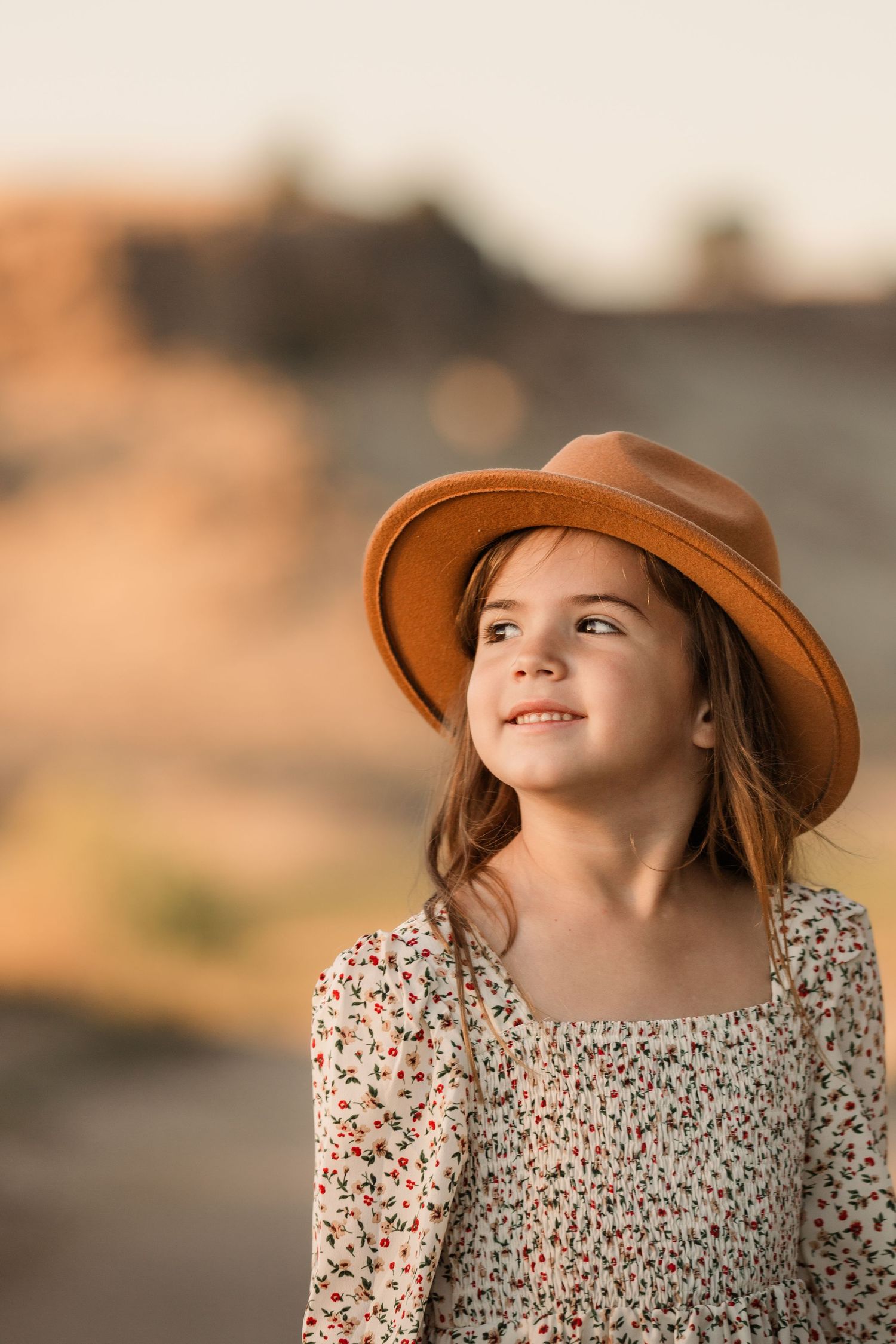 A brown-haired child in a floral dress and wide-brimmed hat gazes upward during golden hour.