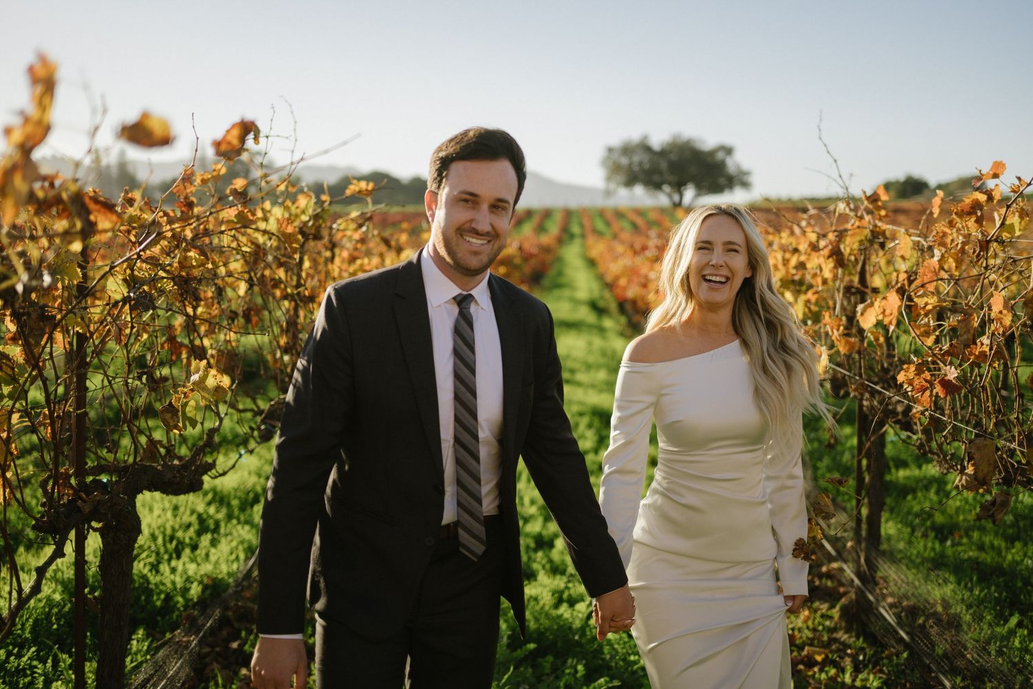 A couple in wedding attire walks together through a scenic vineyard with autumn colored vines in the background.
