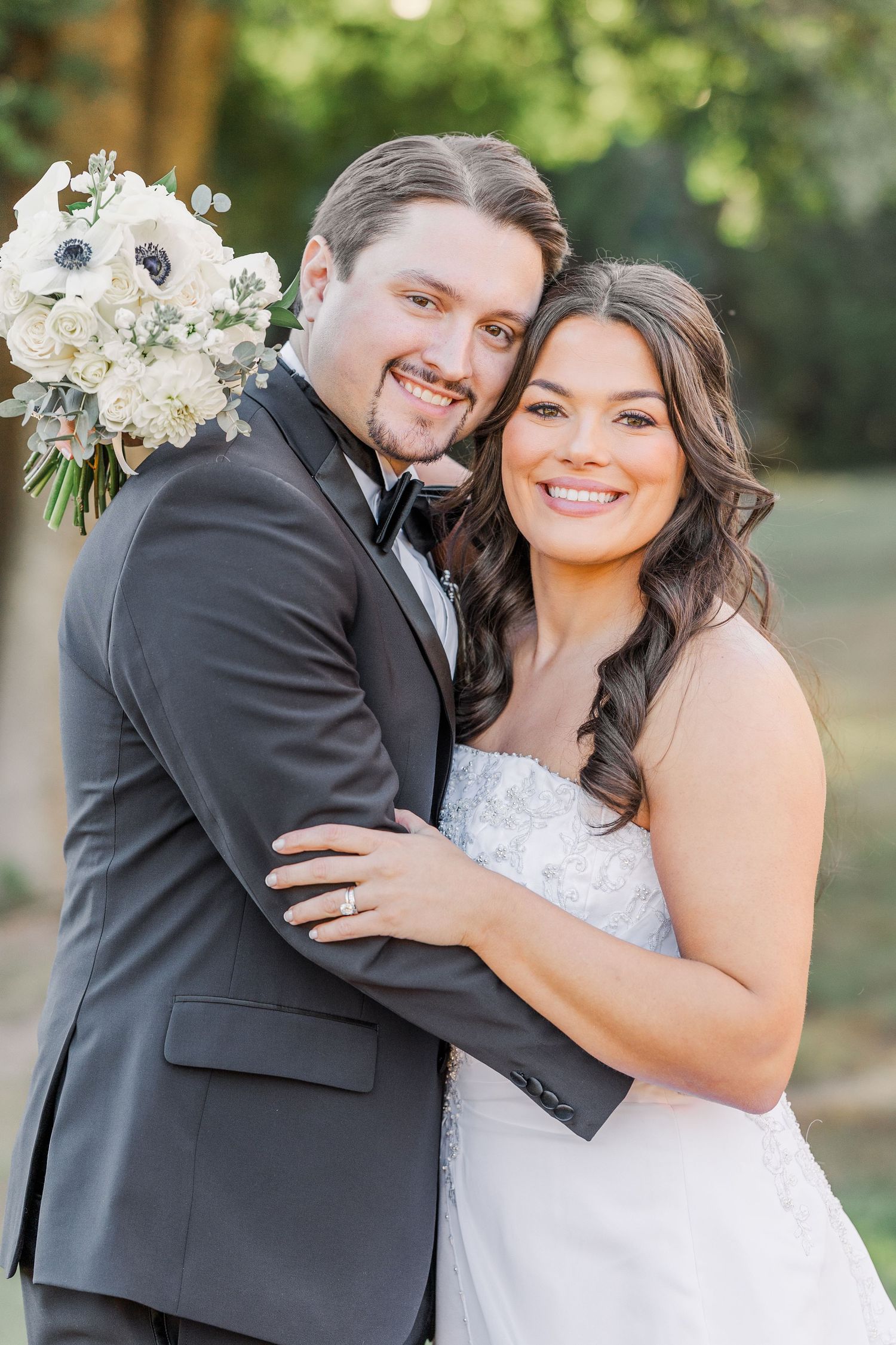 A joyful wedding couple embraces outdoors while wearing traditional black tuxedo and white lace gown.
