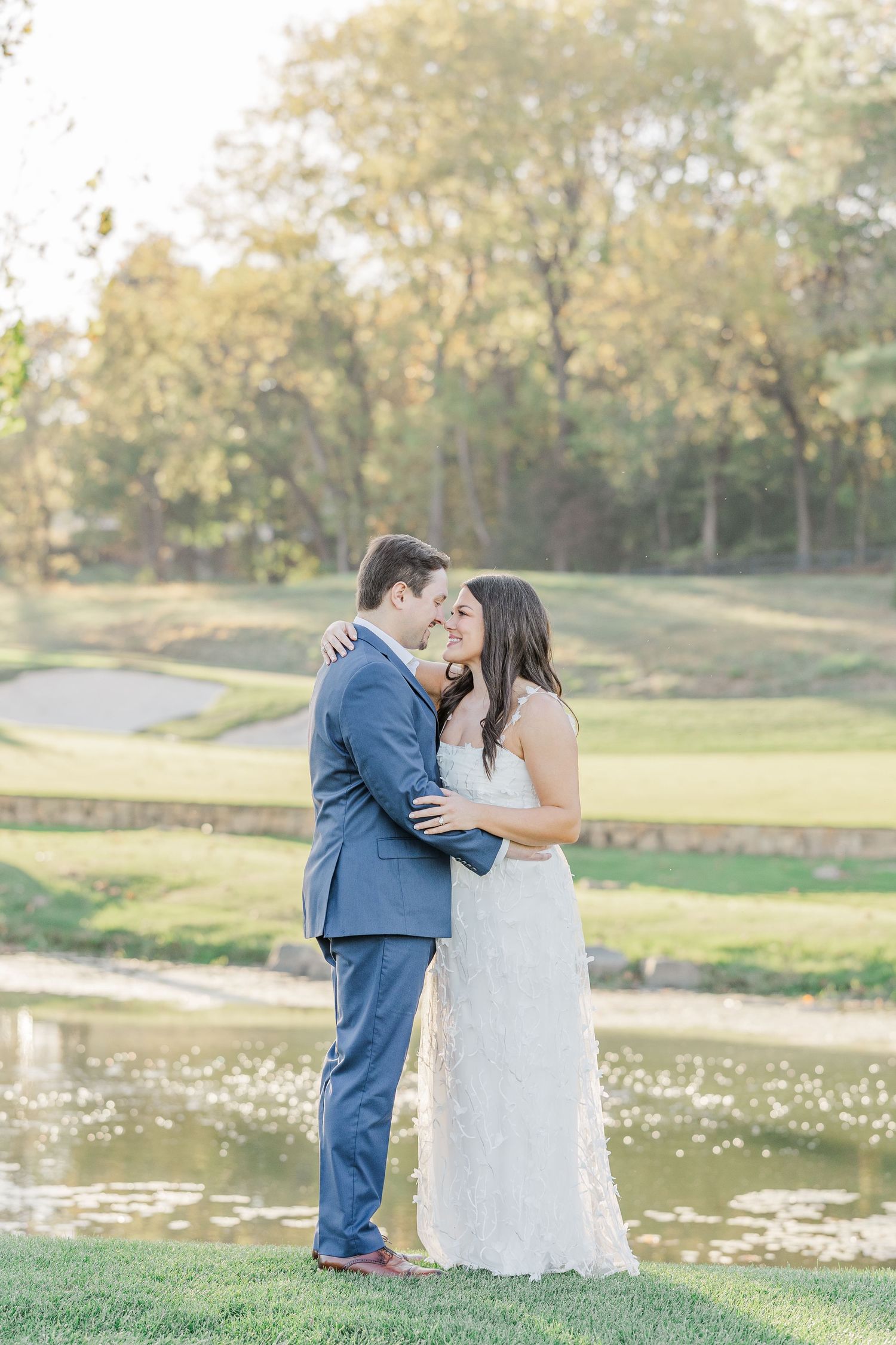 A romantic wedding couple embraces on a golf course with a pond and autumn trees in the background.